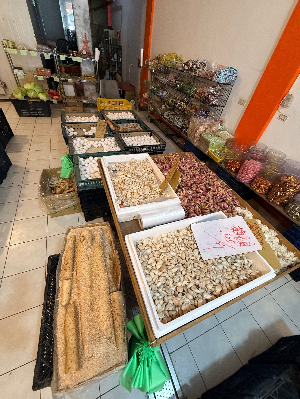 攤位陳列多籃蛋類、蒜頭及乾貨調味料，後方貨架上擺放包裝食品。